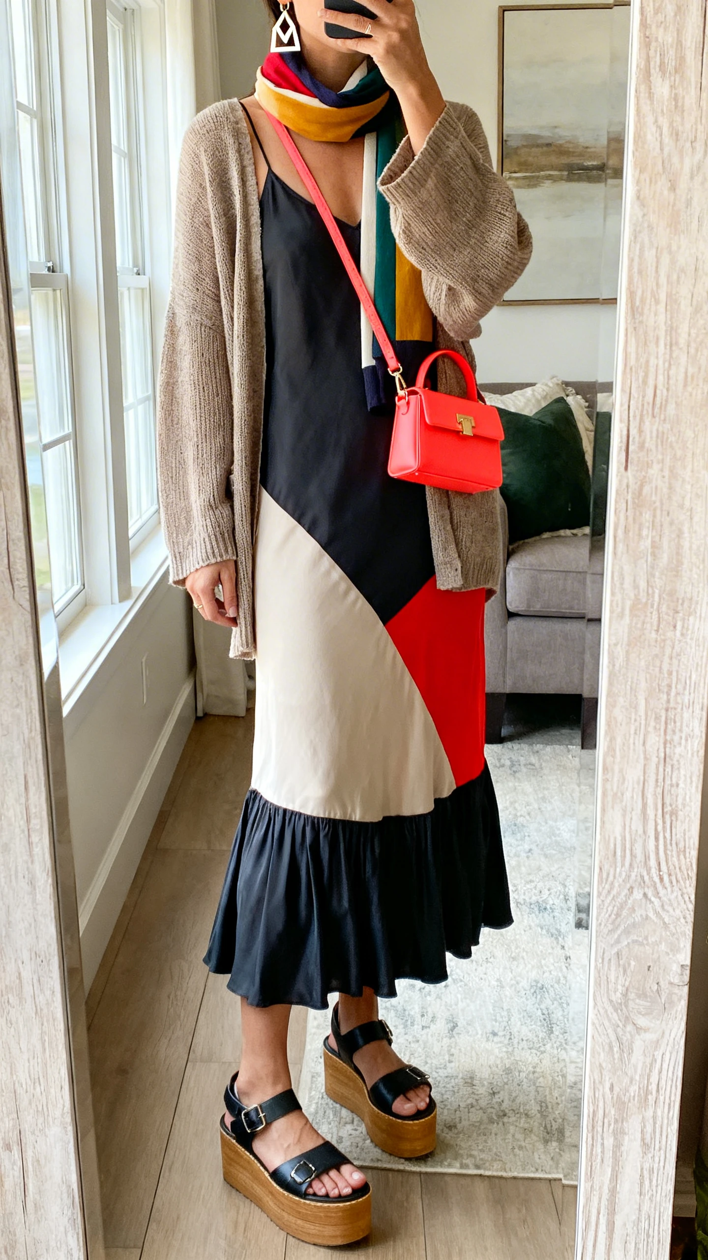 iPhone mirror selfie of a woman wearing an artsy tiered slip midi with a bold color-pop panel, oversized lightweight cardigan, color-block scarf, chunky platform sandals, geometric earrings, and a bright mini shoulder bag, face not visible, cropped at neck, living room full-length mirror, natural window light, iPhone photo quality.