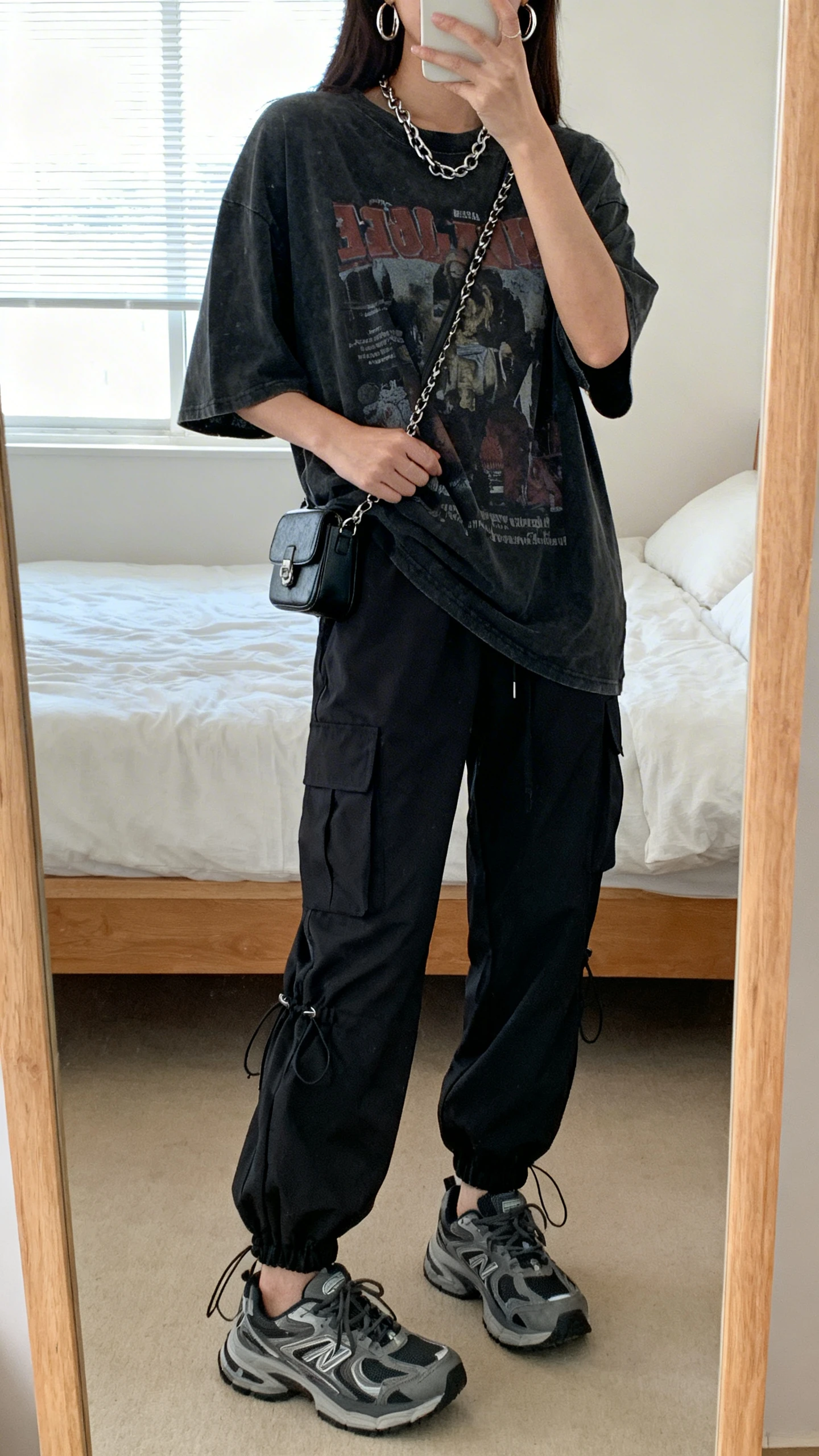 iPhone mirror selfie of a woman wearing black tech cargo pants with cinch hems, an oversized graphic tee, chunky sneakers, a mini crossbody bag, and chain hoops, face not visible, cropped at neck, bedroom mirror, natural window light, iPhone photo quality.