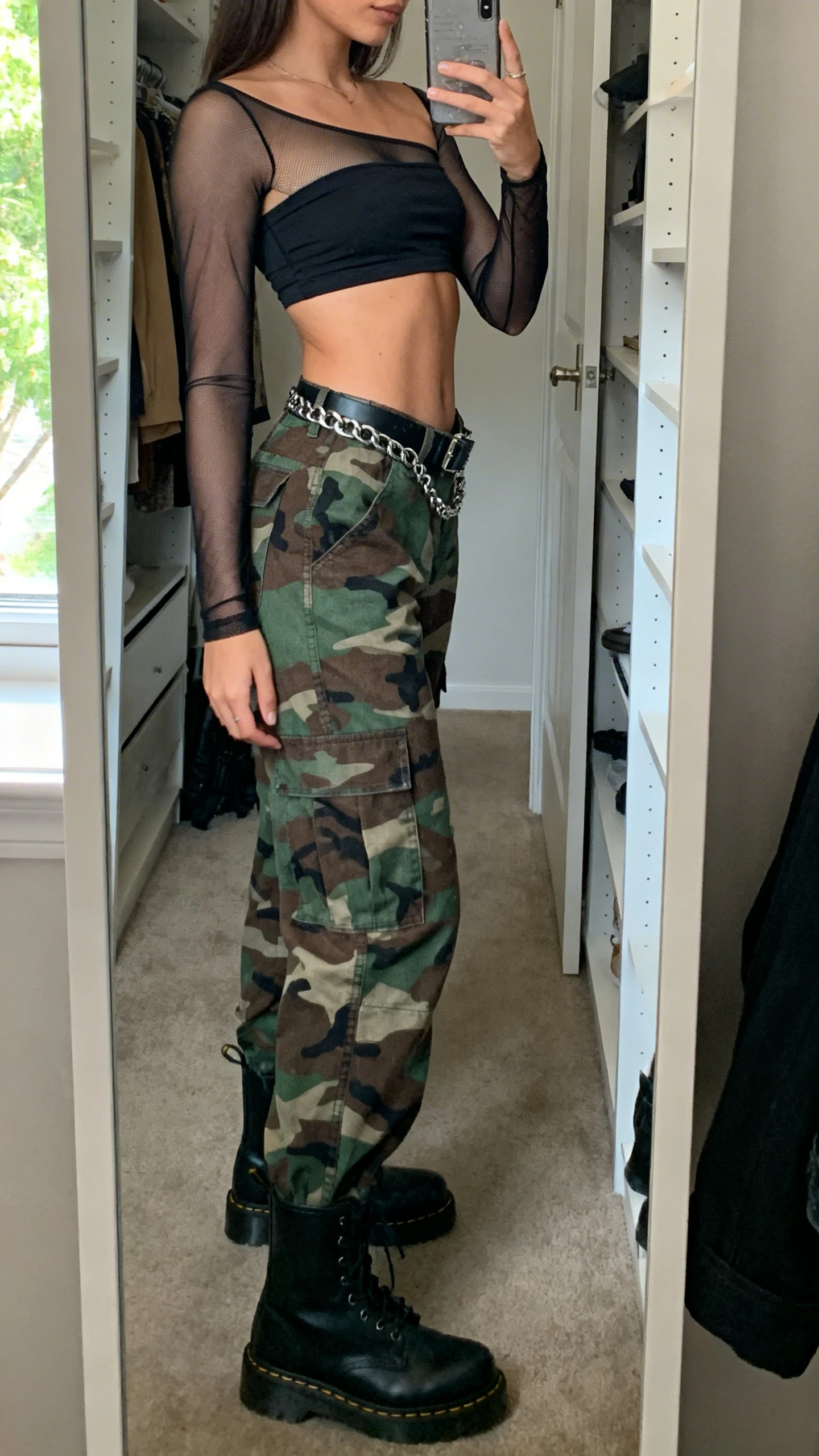 iPhone mirror selfie of a woman wearing camo cargo pants and a tube top crop layered with a sheer mesh long-sleeve, combat boots, chain belt, face not visible, side angle, closet mirror, natural window light, iPhone photo quality.
