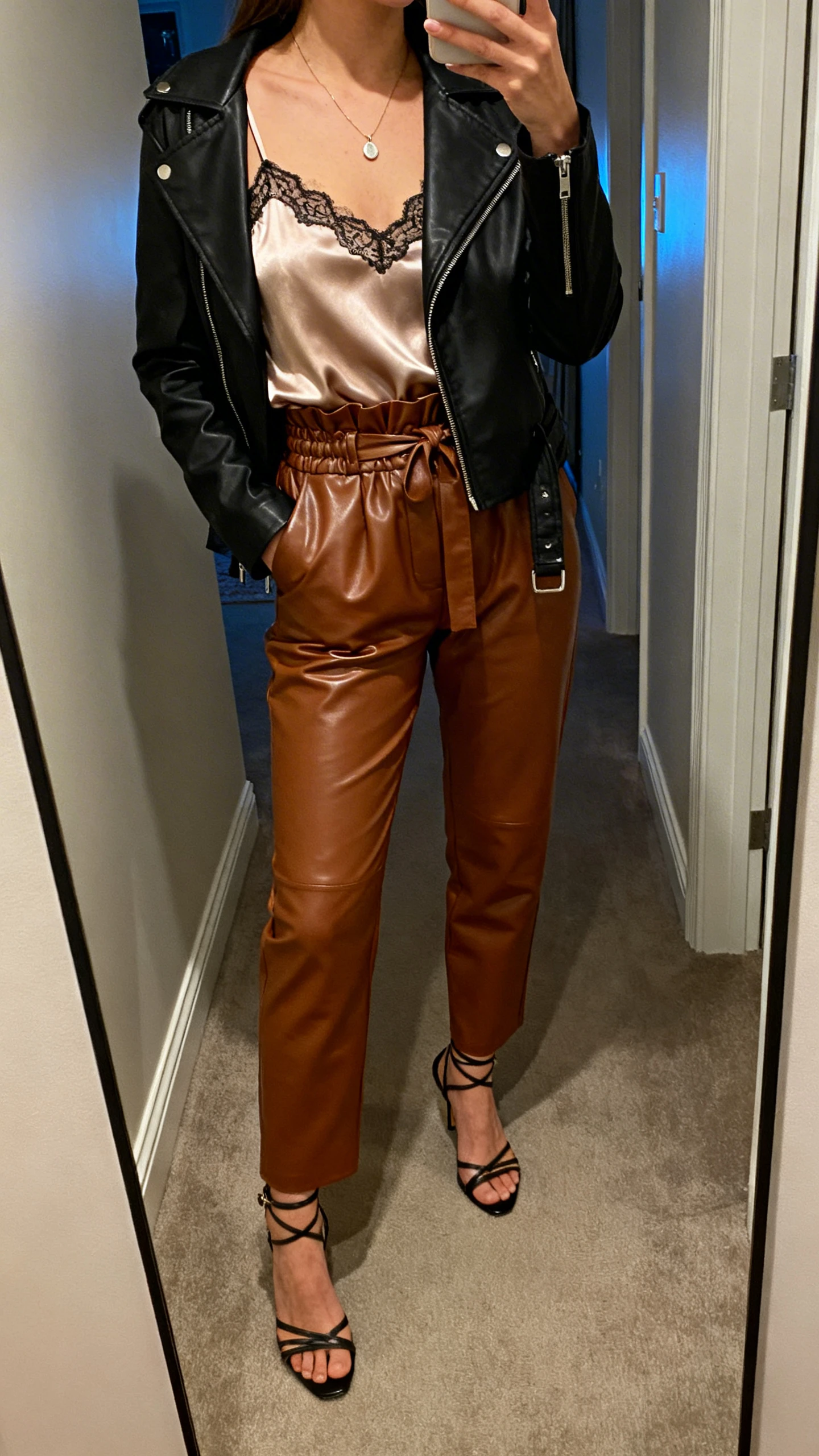 iPhone mirror selfie of a woman wearing paperbag-waist faux leather pants, satin-lace cami tucked in, black moto jacket layered on top, strappy heels, delicate pendant necklace, face not visible, cropped at shoulders, hallway full-length mirror, indoor evening light, iPhone photo quality.