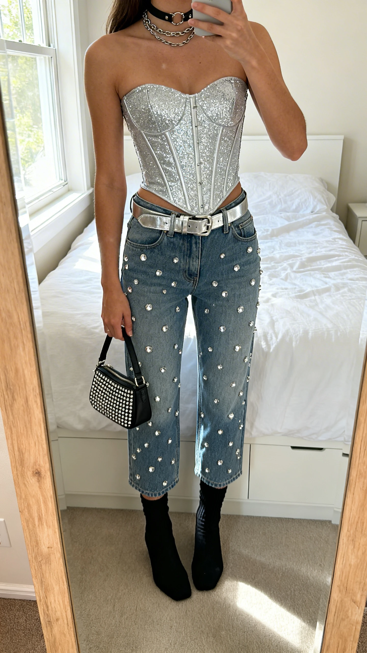 iPhone mirror selfie of a woman wearing rhinestone-studded straight-leg jeans, a silver glitter corset top, optional metallic belt, sleek black sock boots, a choker with short layered chains, and a studded mini bag, face not visible, cropped at shoulders, bedroom mirror, morning window light, iPhone photo quality.