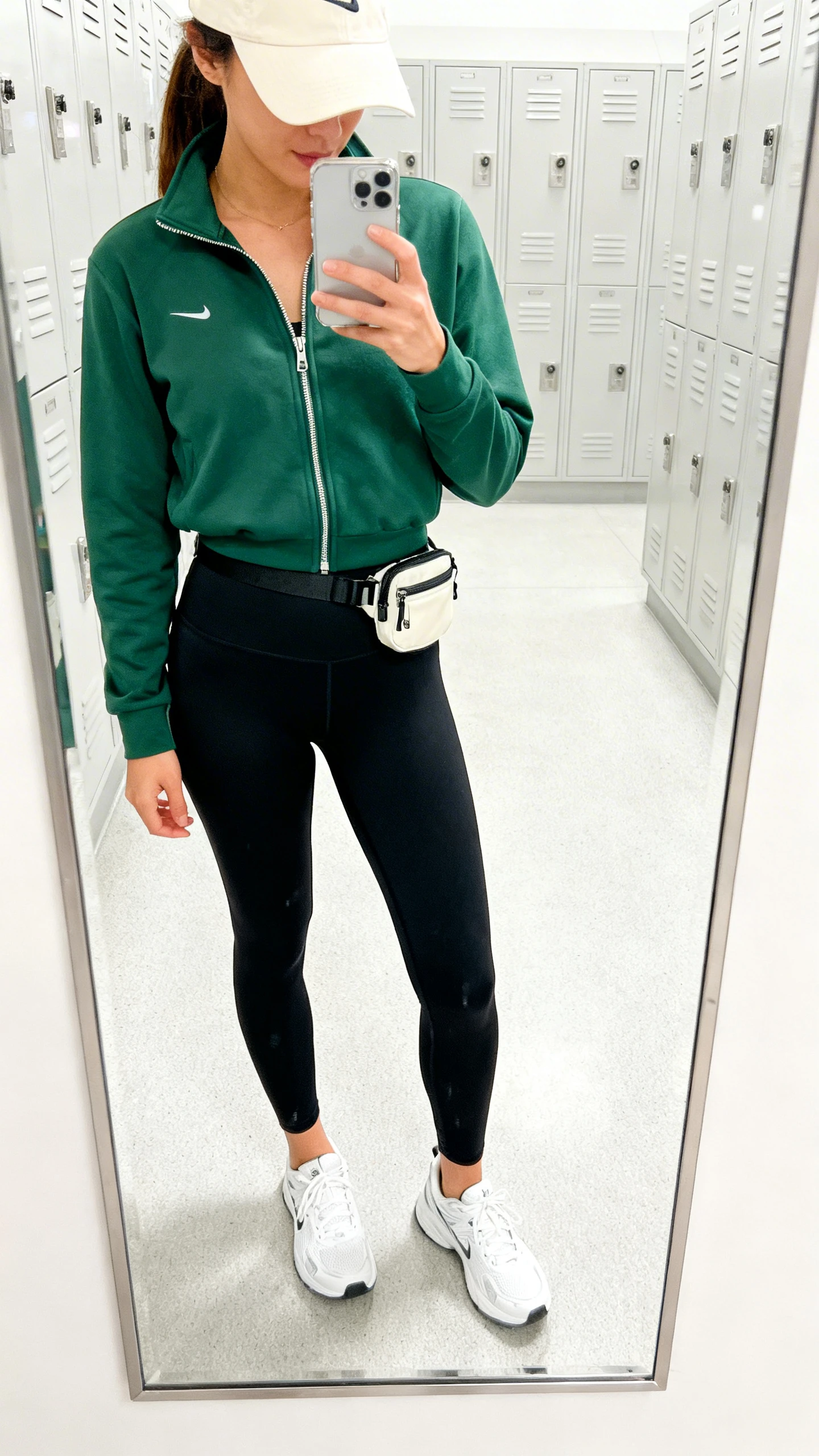 iPhone mirror selfie of a woman wearing sporty-cute athleisure: forest green zip-up track jacket, black high-waist leggings, white running sneakers, a cream baseball cap, and a small belt bag, face not visible, cropped at neck, gym locker-room mirror, bright indoor lighting, iPhone photo quality.