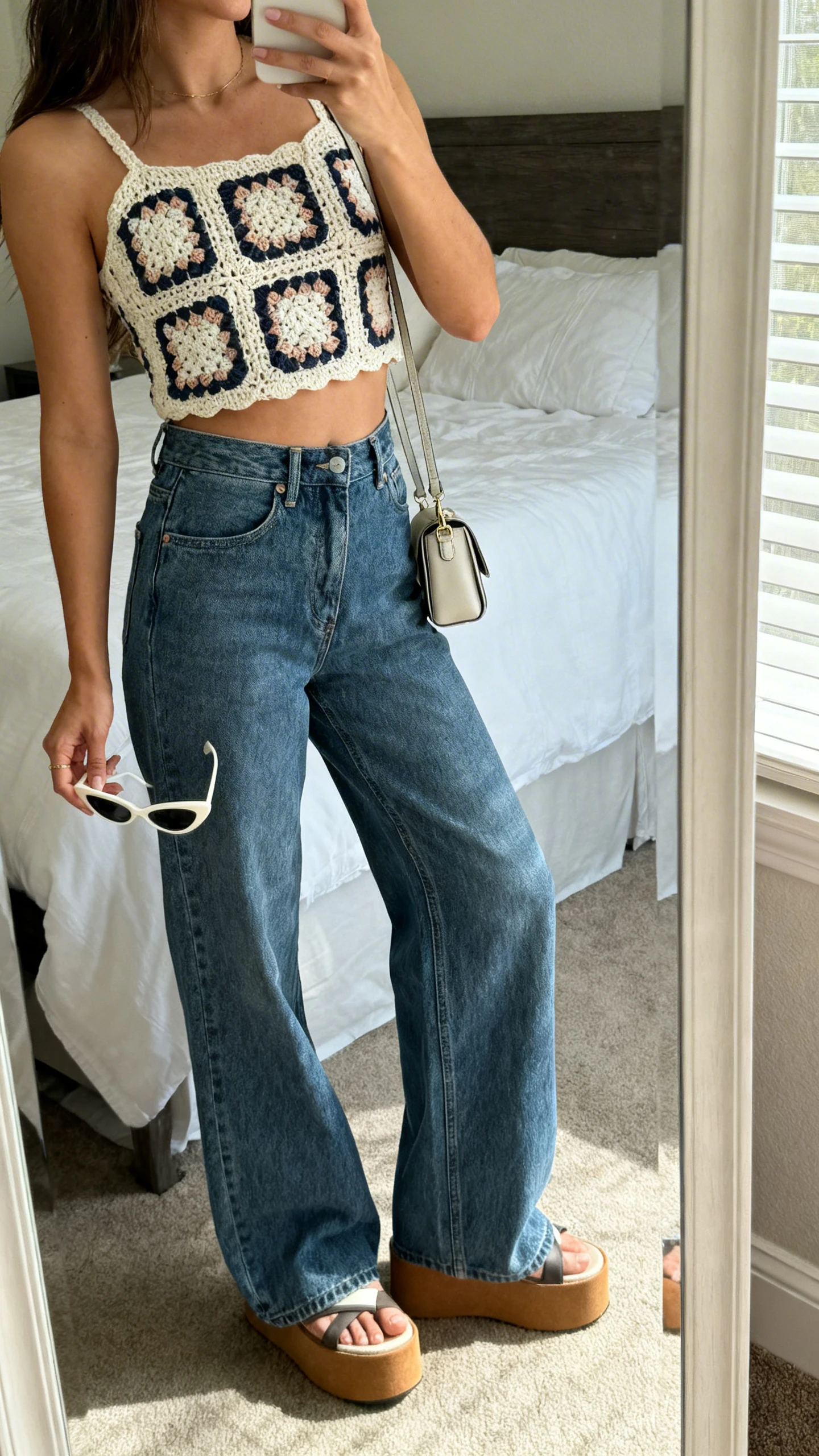 iPhone mirror selfie side angle of a woman wearing a retro granny-square crochet crop top with high-waisted wide-leg jeans, chunky platform sandals, cat-eye sunglasses in hand, and a mini shoulder bag; face not visible, cropped at shoulders, bedroom mirror, natural daylight, iPhone photo quality.