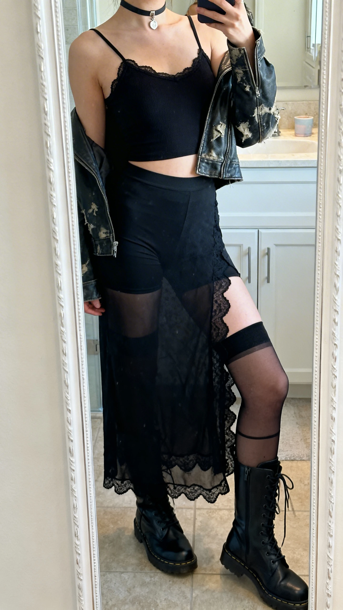 iPhone side-angle mirror selfie of a woman wearing a midi black lace slip dress with scalloped hem over a black jersey bodycon mini or camisole with bike shorts, cropped distressed leather jacket, knee-high lace-up boots, sheer black knee-highs/thigh-high stockings, and a choker with a single charm, face not visible, bathroom mirror, soft indoor lighting, iPhone photo quality.