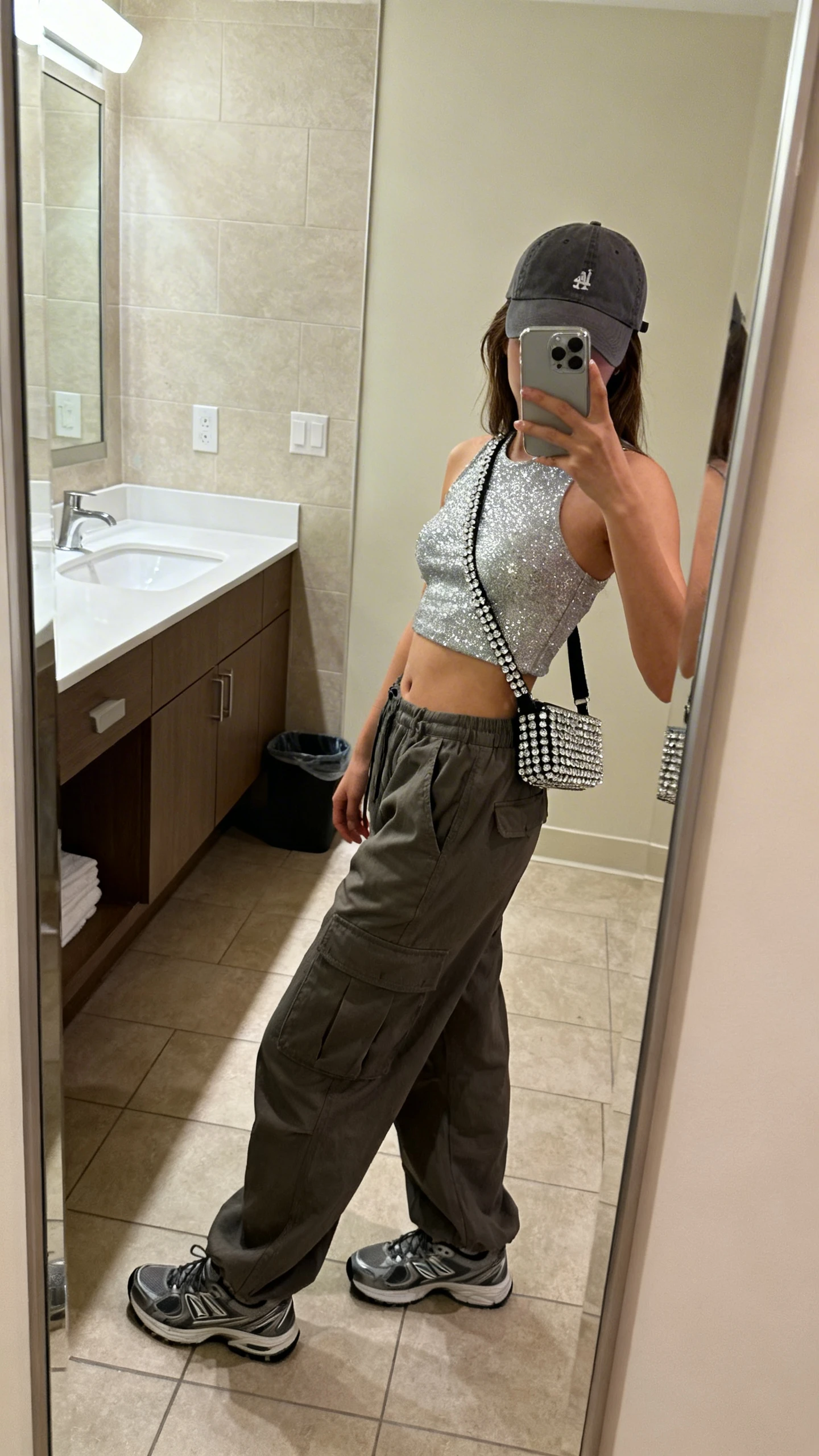 iPhone side-angle mirror selfie of a woman wearing a glitter crop top, slouchy cargo pants, sleek trainers, baseball cap, and rhinestone-accent crossbody; face not visible, bathroom full-length mirror, soft indoor lighting, iPhone photo quality.