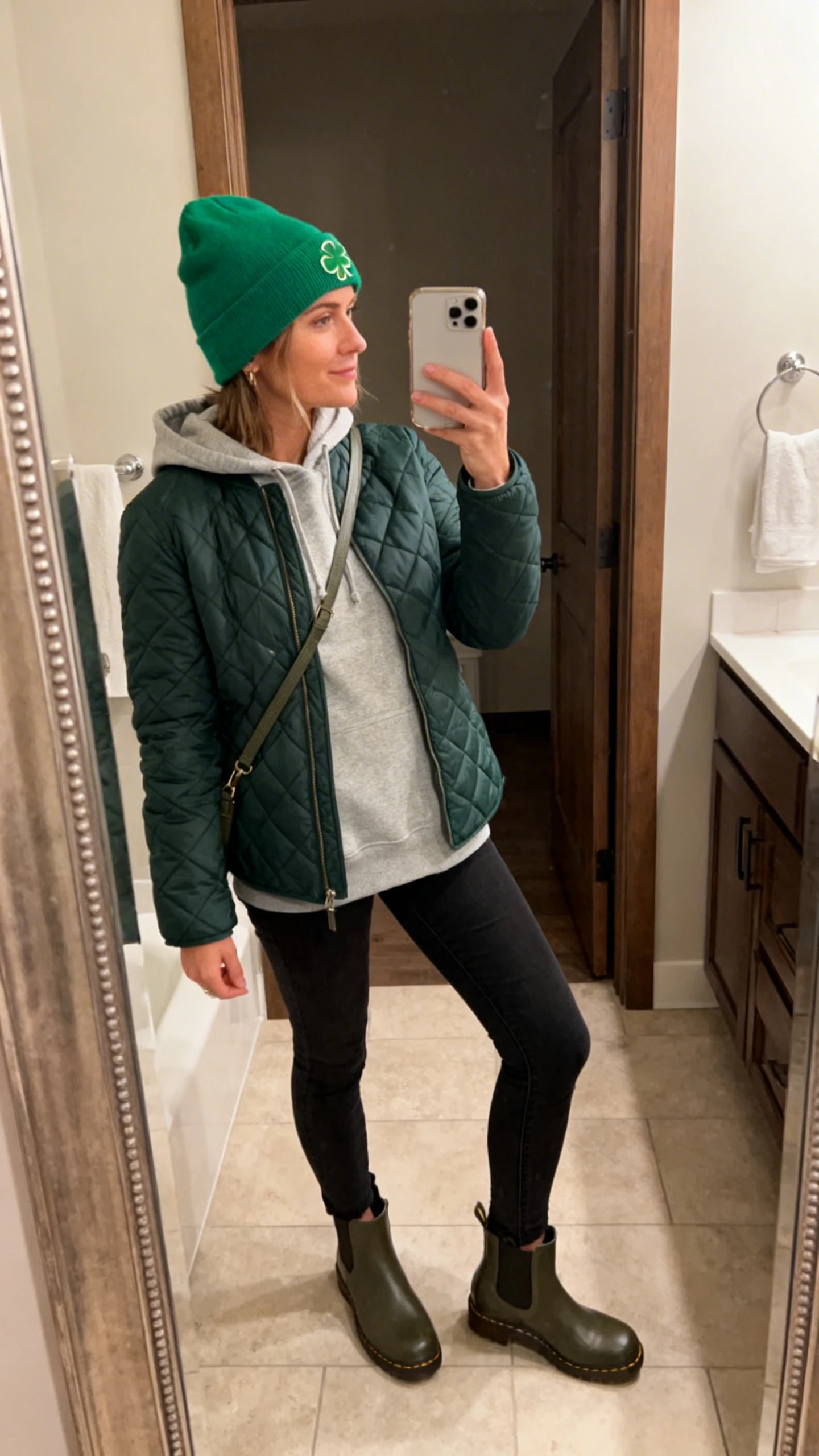 iPhone side-angle mirror selfie of a woman wearing a cozy layered look: heather grey hoodie under a dark green quilted jacket, black skinny jeans, waterproof Chelsea boots, a shamrock-green beanie, and a crossbody bag, face not visible, bathroom mirror, warm indoor lighting, iPhone photo quality.