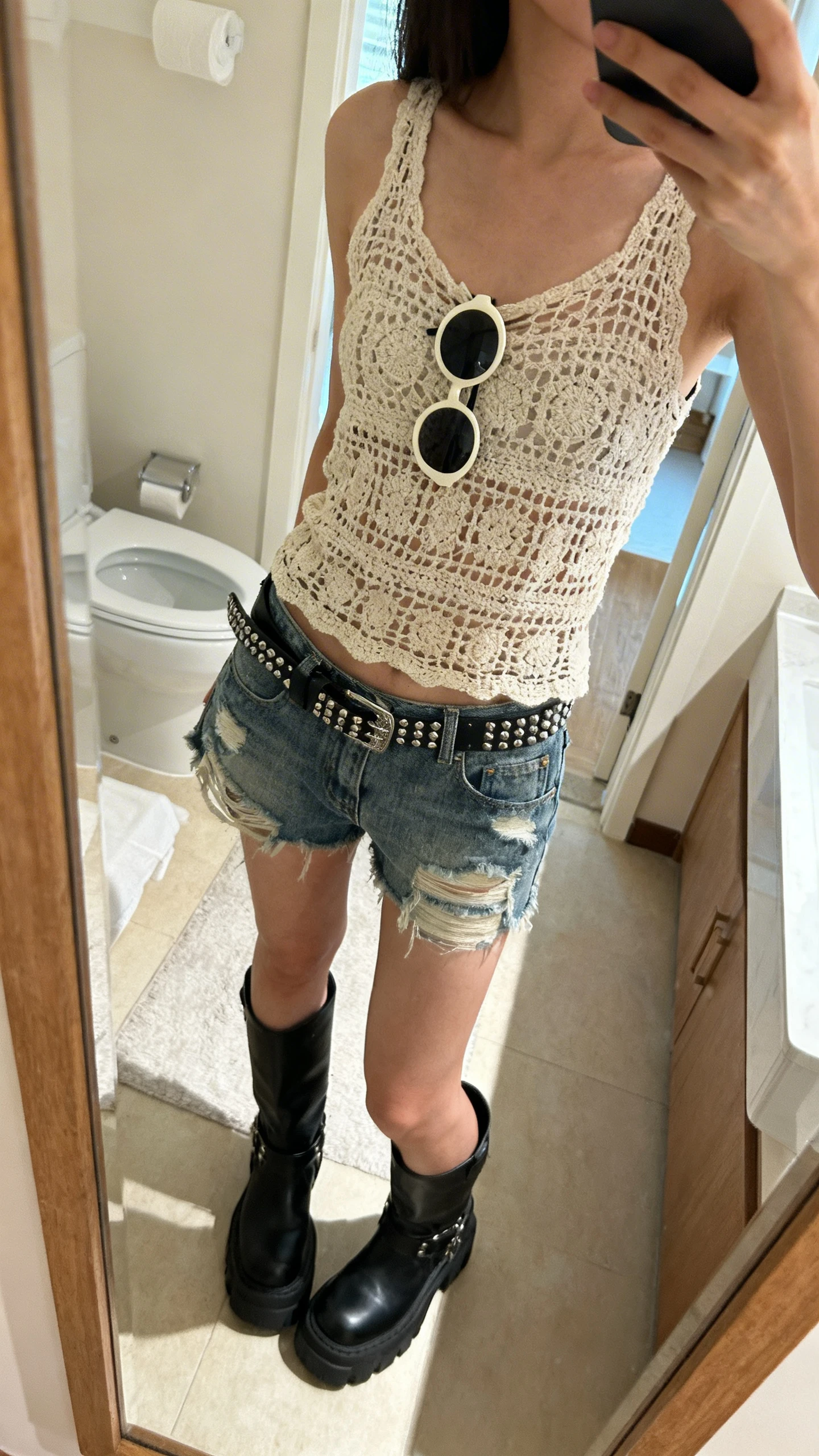 iPhone side-angle mirror selfie of a woman wearing a retro-grunge crochet tank with distressed denim shorts, chunky black boots, a studded belt, and round sunglasses hooked on the tank, face not visible, bathroom mirror, indoor morning light, iPhone photo quality.