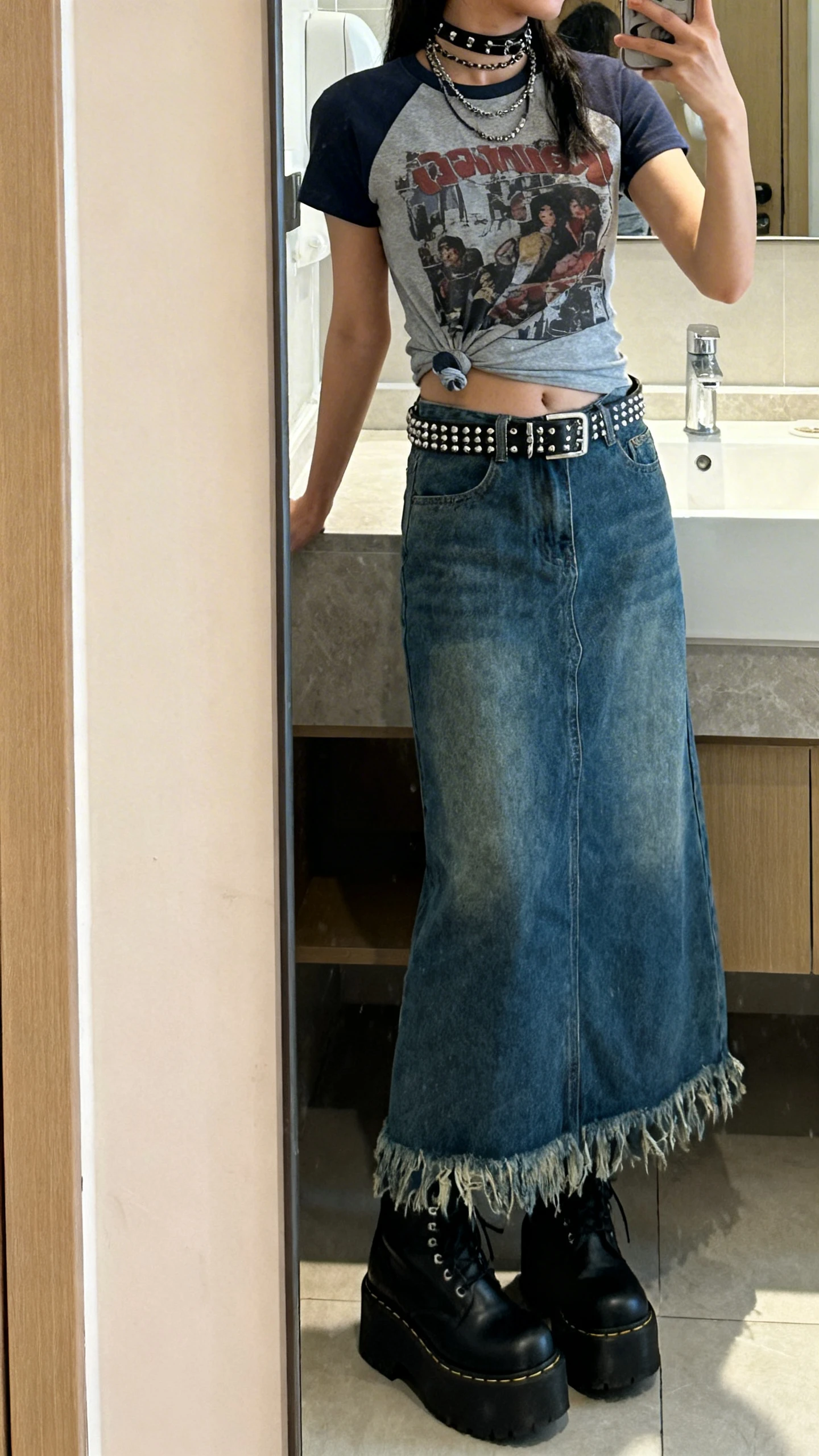 iPhone side-angle mirror selfie of a woman wearing a denim maxi skirt with raw edges, graphic tee tucked in, platform boots, studded belt, and layered chokers, face not visible, bathroom mirror, soft indoor lighting, iPhone photo quality.