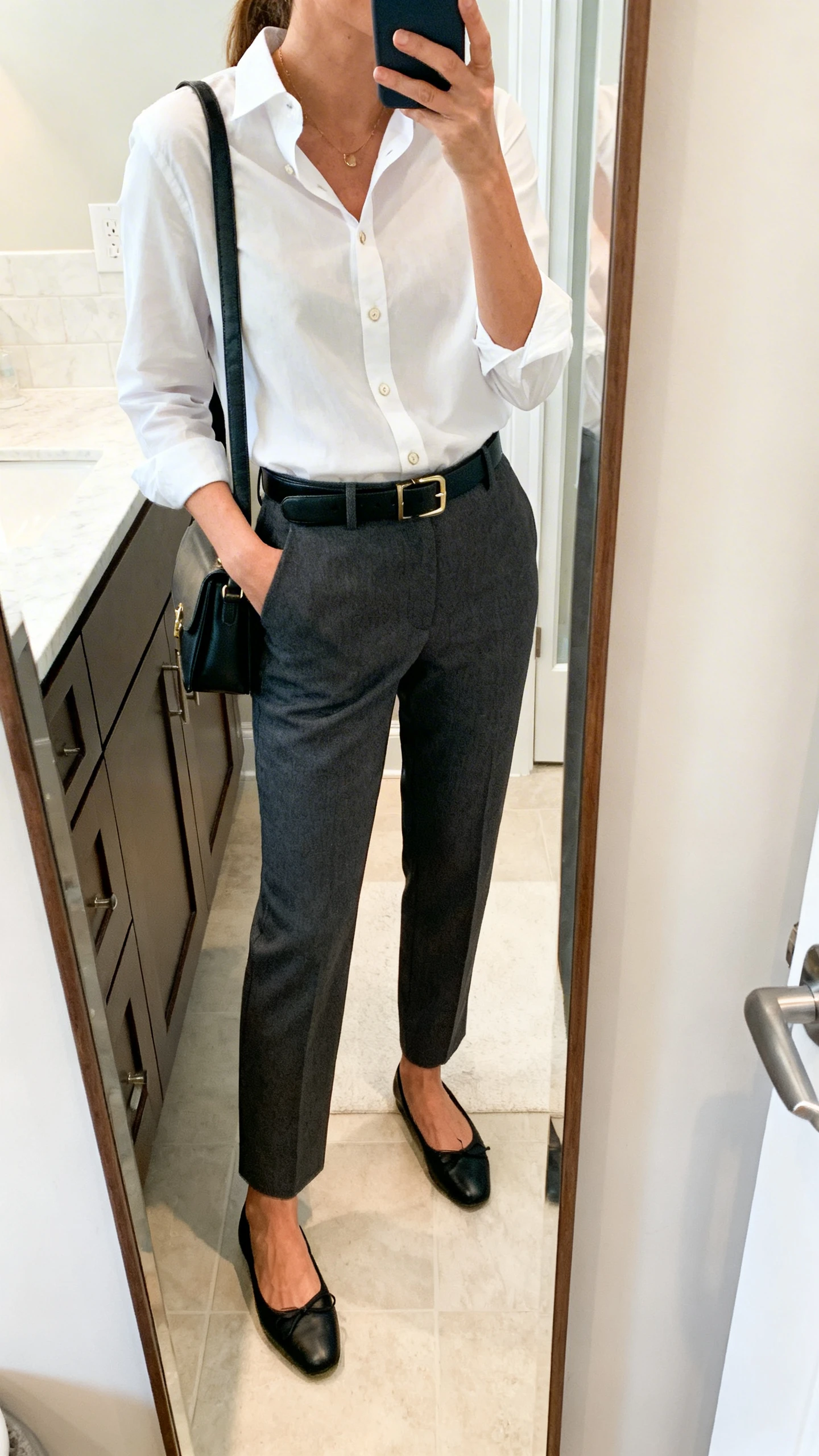 iPhone side-angle mirror selfie of a woman wearing tailored charcoal trousers with a crisp white button-up, black ballet flats, a leather belt, and a structured shoulder bag, face not visible, bathroom full-length mirror, soft indoor lighting, iPhone photo quality.
