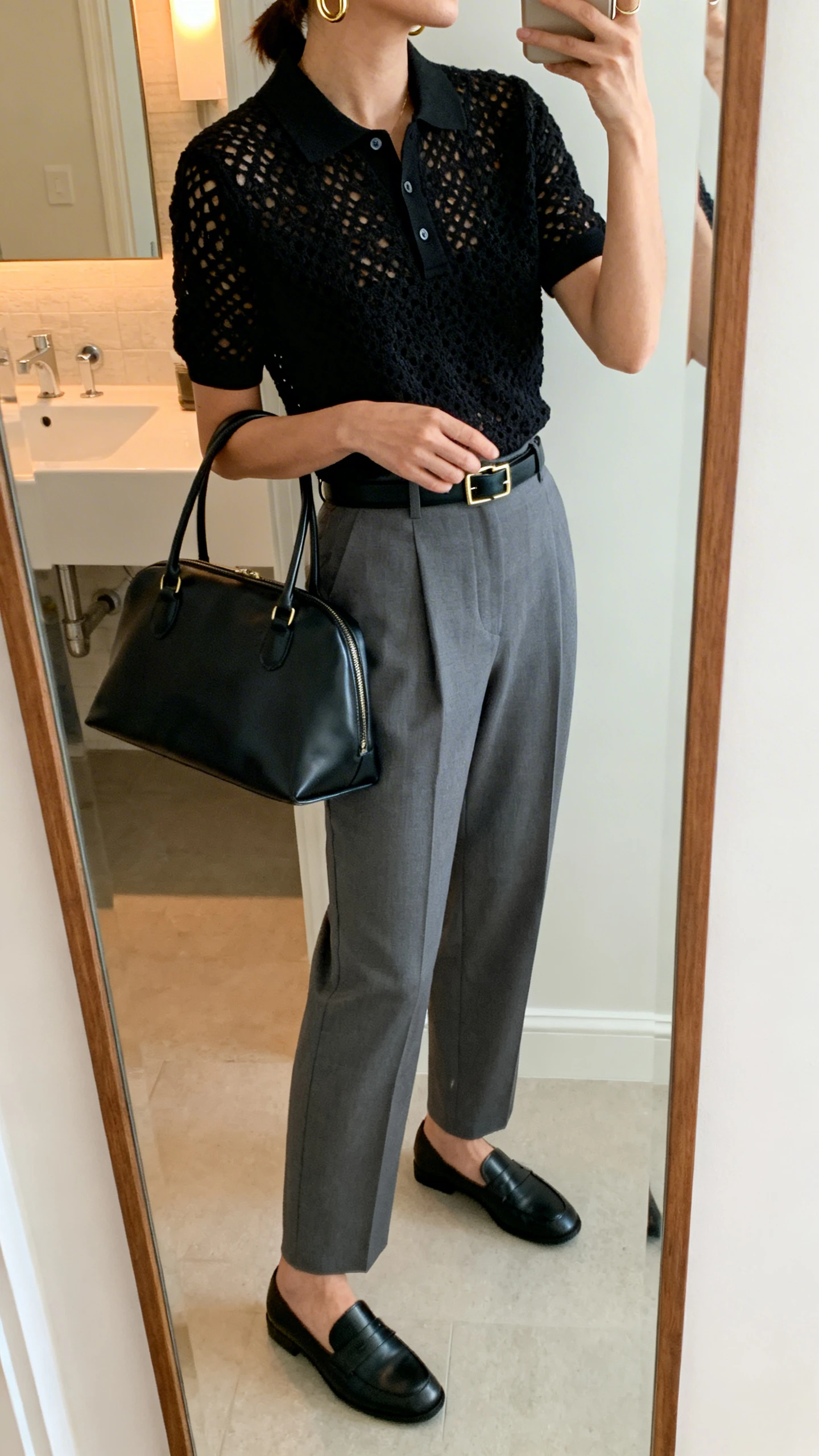 iPhone side-angle mirror selfie of a woman wearing a black crochet polo top, high-waisted tailored gray trousers, sleek black loafers, minimal gold hoops, a structured black leather tote, and a slim black belt, face not visible, cropped at shoulders, bathroom mirror with soft indoor lighting, iPhone photo quality.