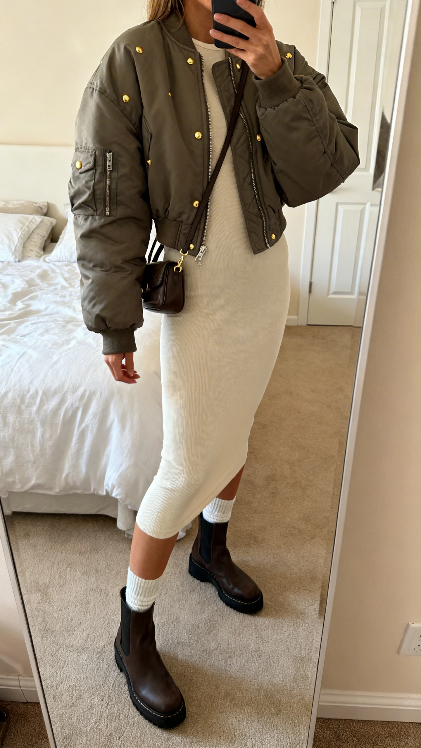 iPhone side-angle mirror selfie of a woman wearing a minimalist bodycon dress with an oversized bomber jacket, sleek sock boots, tiny shoulder bag, and subtle gold studs, face not visible, cropped at shoulders, bedroom mirror, warm indoor lighting, iPhone photo quality.