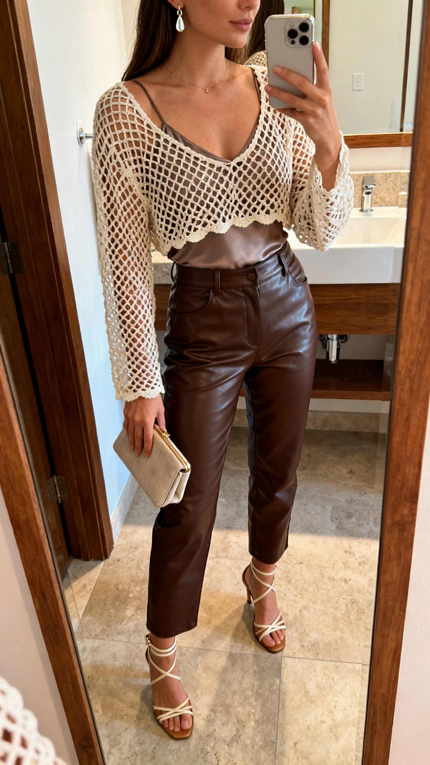 iPhone side-angle mirror selfie of a woman wearing a crochet mesh top over a sleek camisole, high-waisted leather pants, strappy heels, small clutch, and delicate drop earrings; face not visible, bathroom mirror, warm indoor lighting, iPhone photo quality.