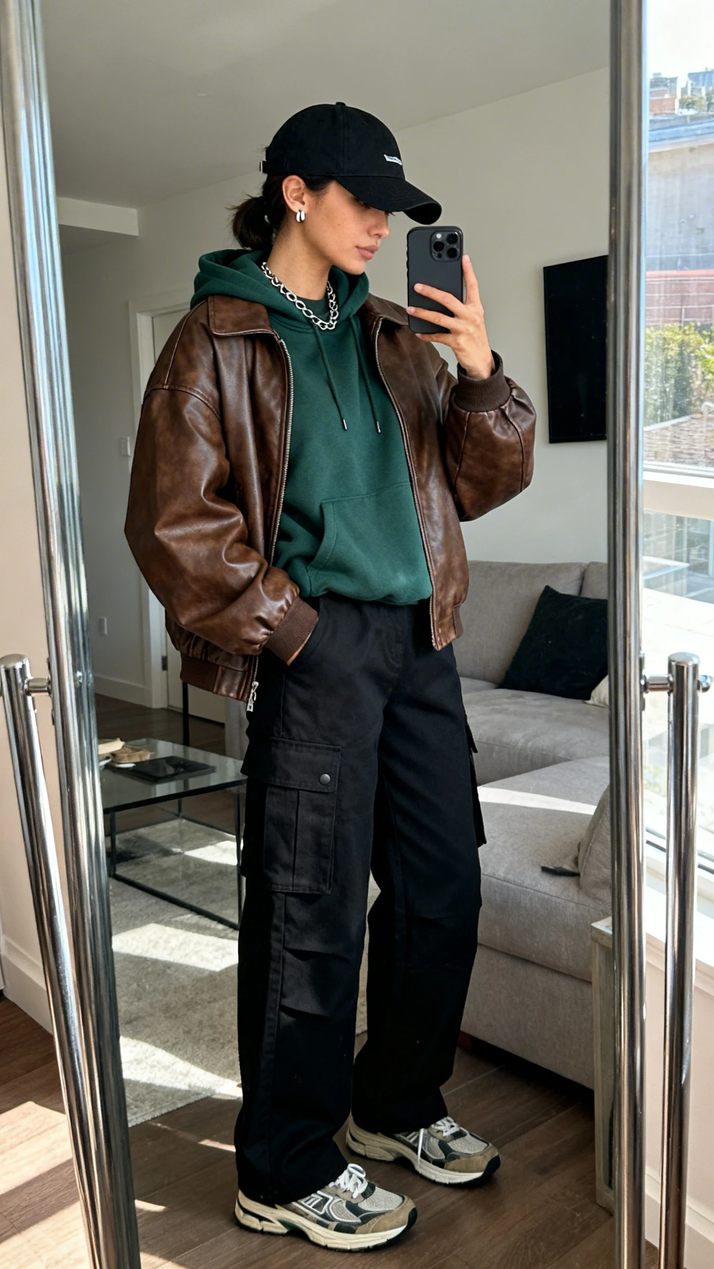 iPhone side-angle mirror selfie of a woman wearing a relaxed brown leather bomber, boxy forest green hoodie, high-waisted black cargo pants, retro sneakers, a black baseball cap, minimal chain necklace and stud earrings, face not visible, living room full-length mirror with natural daylight, iPhone photo quality.