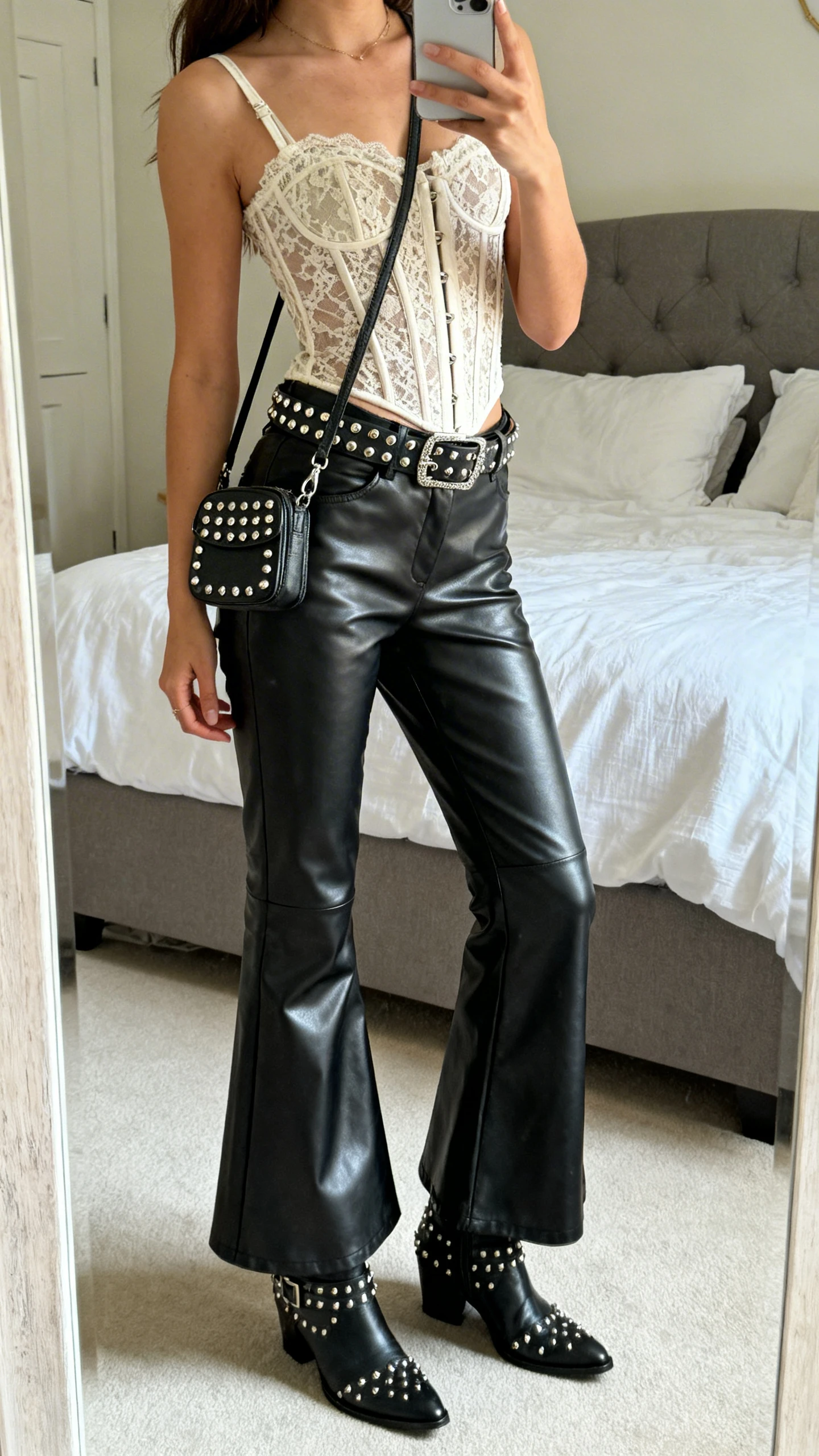 iPhone side-angle mirror selfie of a woman wearing flared faux leather pants, corset-style lace top, studded belt and studded ankle boots, small crossbody with metal studs, face not visible, bedroom mirror, soft natural daylight, iPhone photo quality.