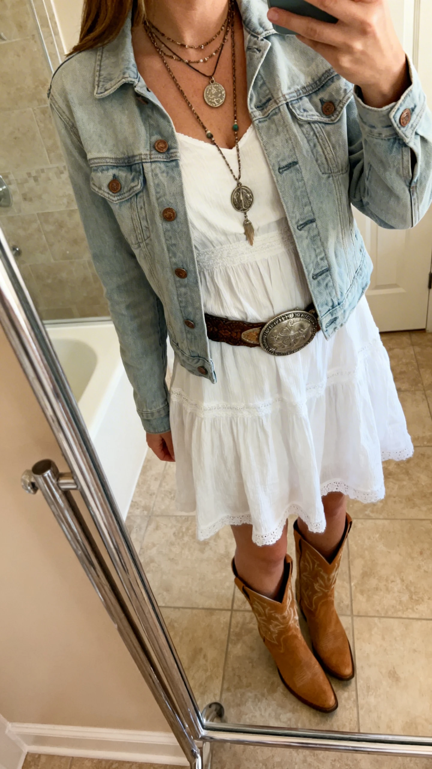 iPhone side-angle mirror selfie of a woman wearing a white sundress layered with a light denim jacket, layered pendant necklaces, concho belt, and tan cowboy boots, face not visible, cropped at neck, bathroom mirror, warm indoor lighting, iPhone photo quality.