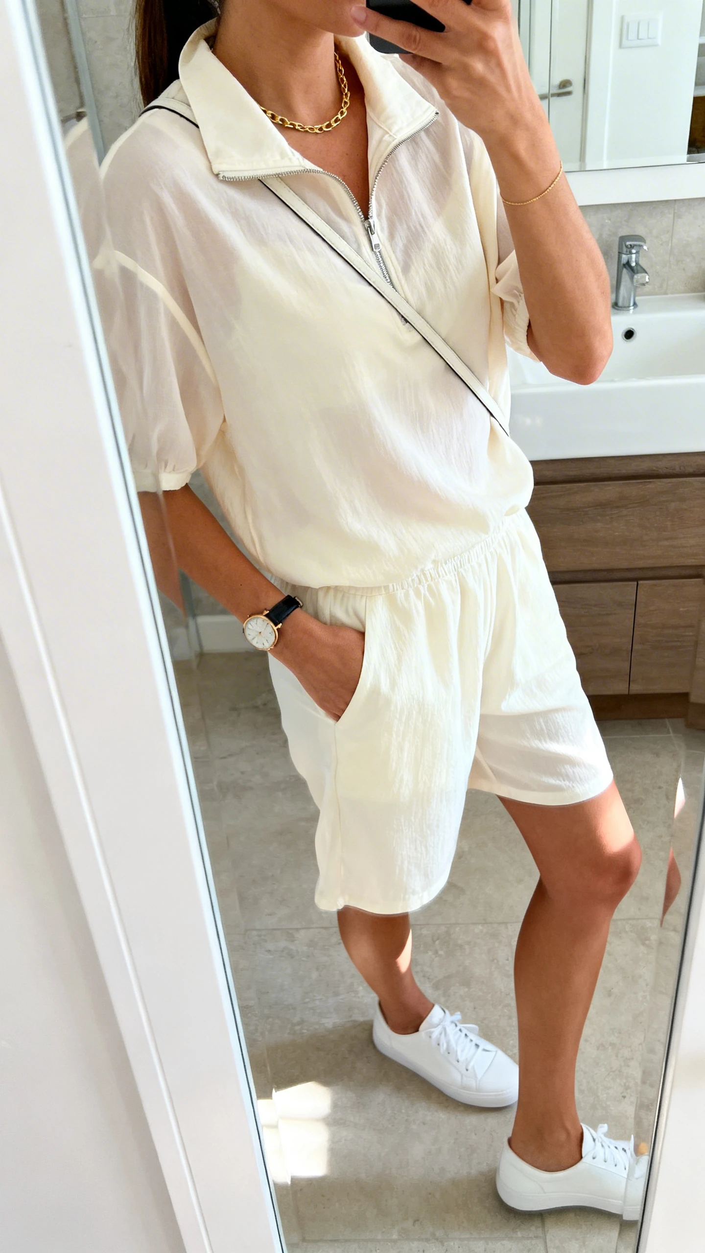 iPhone side-angle mirror selfie of a woman wearing an airy collared set (lightweight collared zip top and relaxed shorts) with sleek sport-luxe minimalism: white low-profile sneakers, thin gold chain, simple watch, and a clean crossbody, face not visible, bathroom mirror, bright natural light, iPhone photo quality.