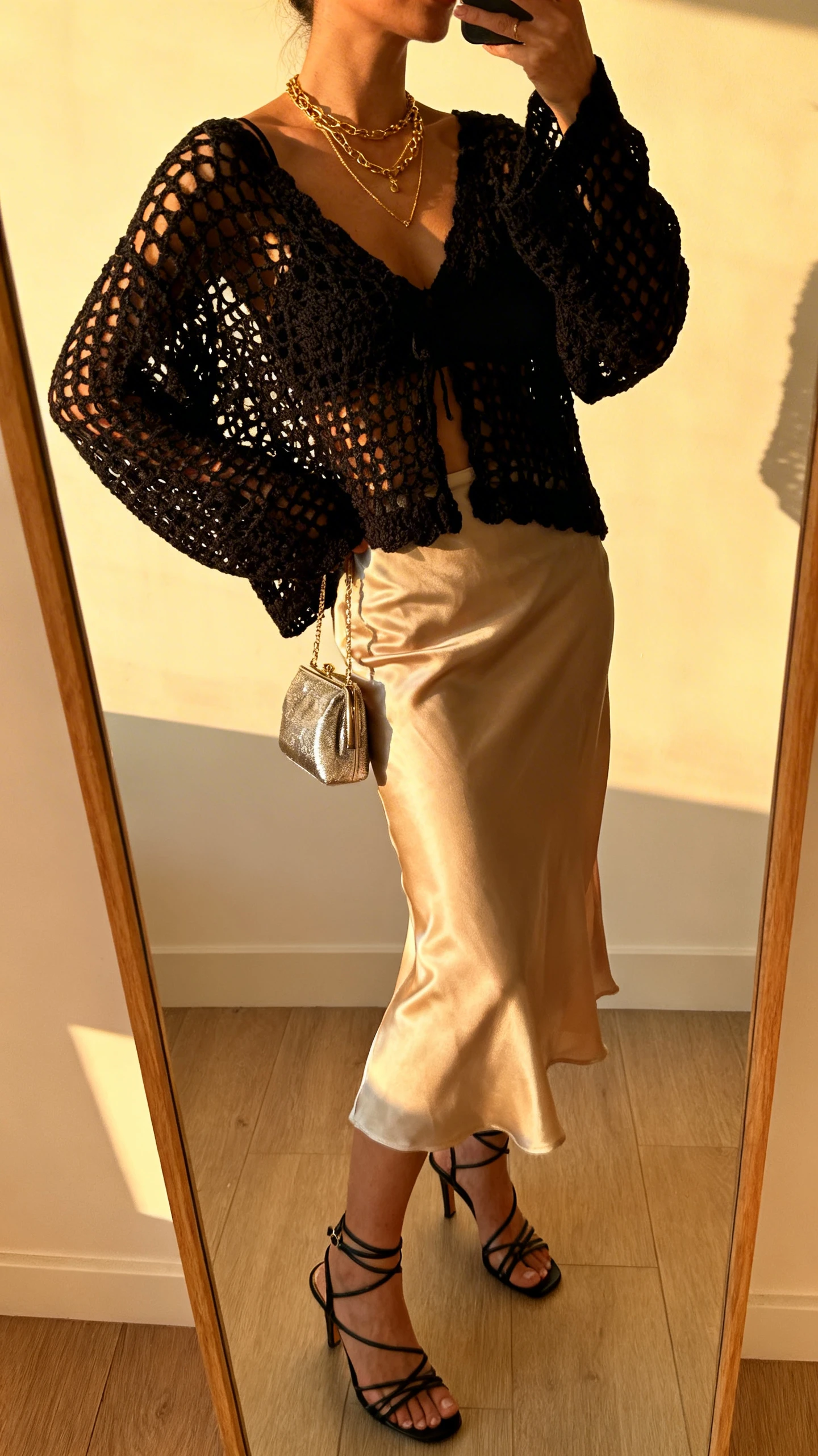 iPhone side-angle mirror selfie of a woman wearing a black open-knit crochet top over a silky champagne slip skirt, strappy heeled sandals, delicate layered gold necklaces, and a small metallic evening bag, face not visible, cropped at shoulders, warm indoor lighting by a window at golden hour, iPhone photo quality.